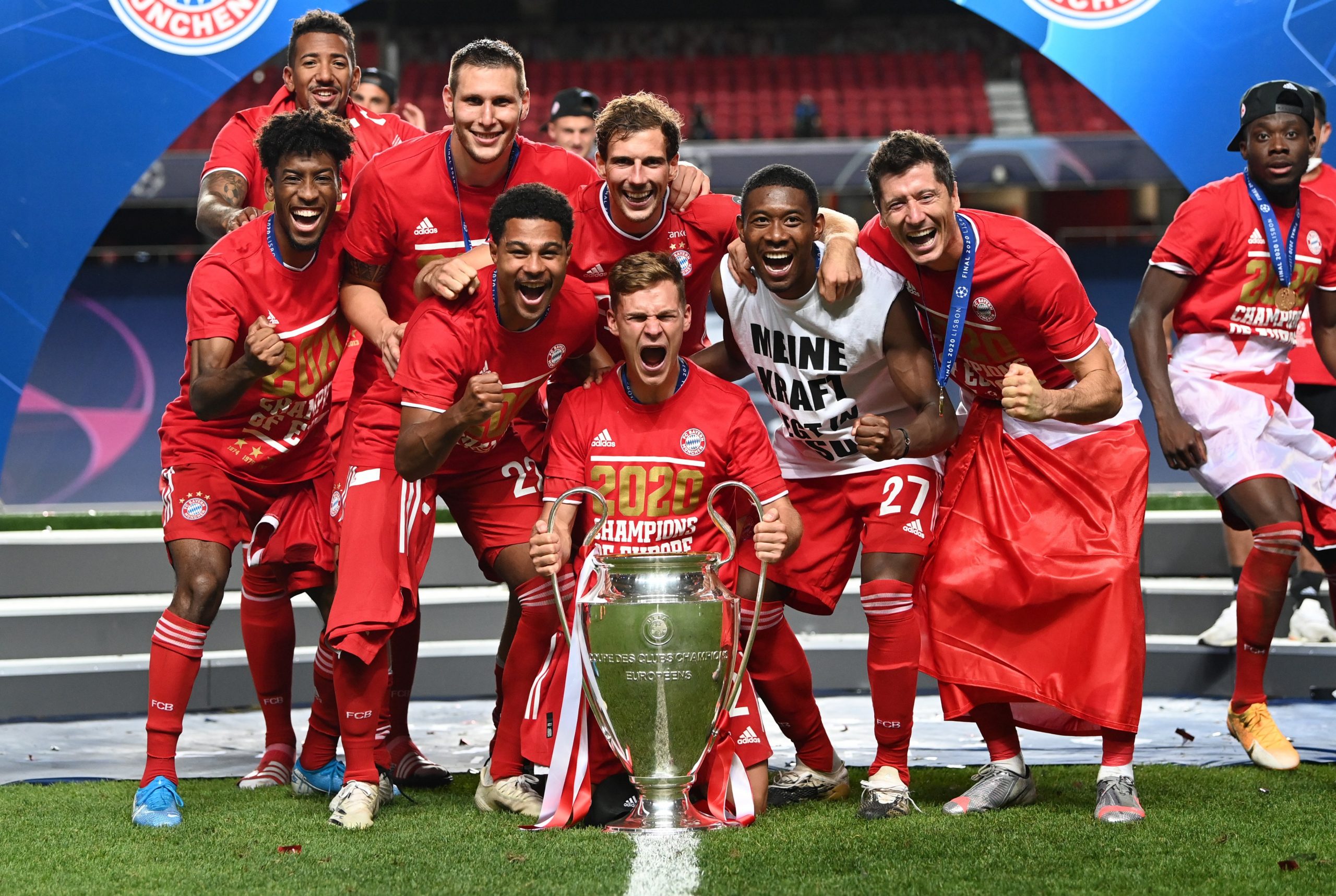  The image shows Bayern Munich players celebrating their victory in the 2019-20 UEFA Champions League with a trophy in the center and some players on their knees while others are standing with smiles on their faces.